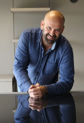 Photographie d'un conseiller en agencement intérieur professionnel, âgé d'environ 45-55 ans, vêtu d'une tenue business casual élégante (chemise ou blazer de couleur neutre), debout dans un showroom moderne et lumineux d'agencement haut de gamme. Il doit regarder directement vers l'appareil photo avec une expression accueillante et professionnelle, légèrement souriant. En arrière-plan flou, on distingue des éléments d'exposition : échantillons de carrelage premium, revêtements de sol, vitrines de mobilier de salle de bain design. L'ambiance doit être chaleureuse, confiante et haut de gamme, reflétant l'expertise et l'approche personnalisée du service. Lumière naturelle ou professionnelle créant une atmosphère de confiance et de professionnalisme.